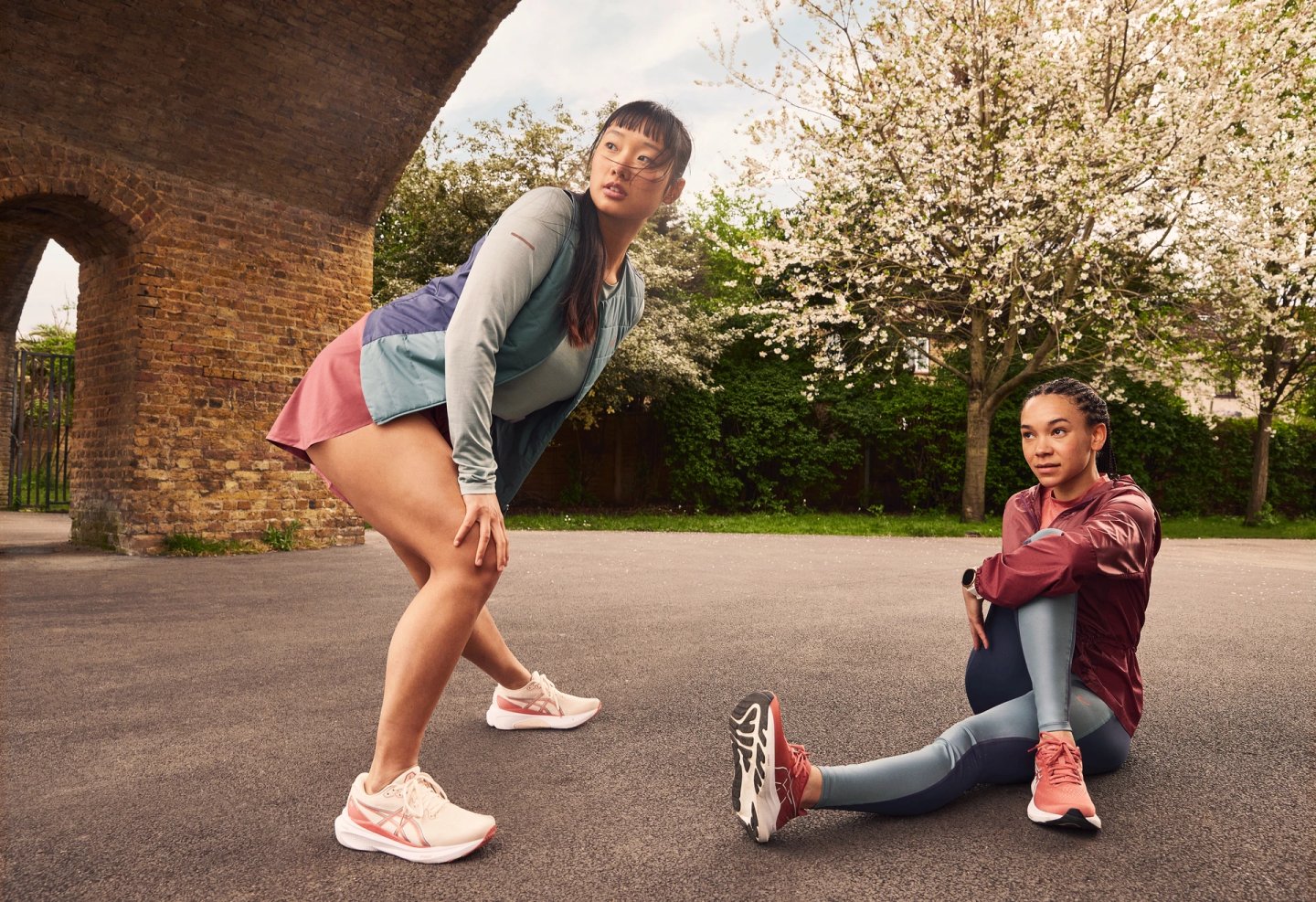 Two women in ASICS running shoes stretching in a park, one standing bending forward, one seated on ground, spring trees behind.