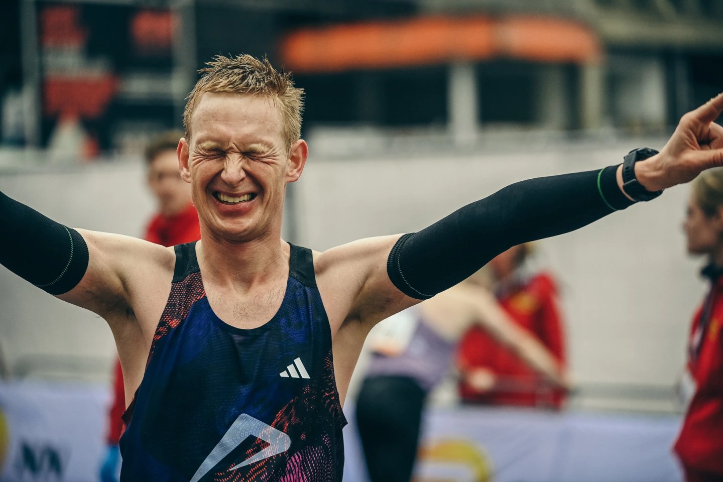 Male runner celebrates at race finish line, arms wide open, wearing dark blue Adidas singlet and black compression arm sleeves.