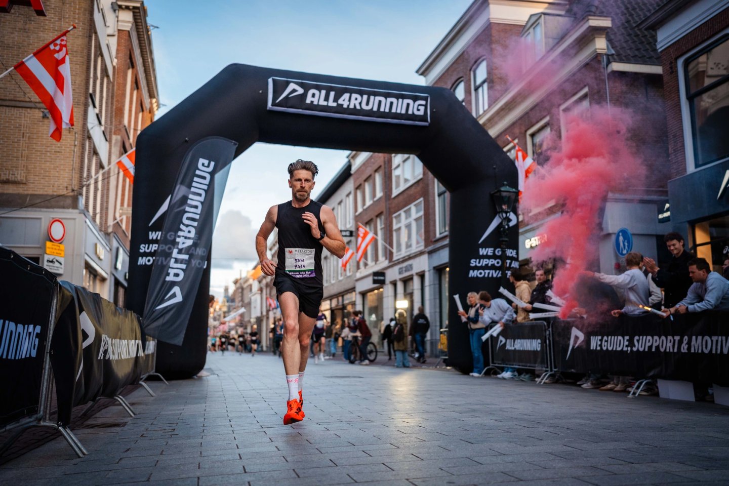Male runner in black vest and orange shoes crossing finish line arch on cobblestone city street with red smoke and spectators