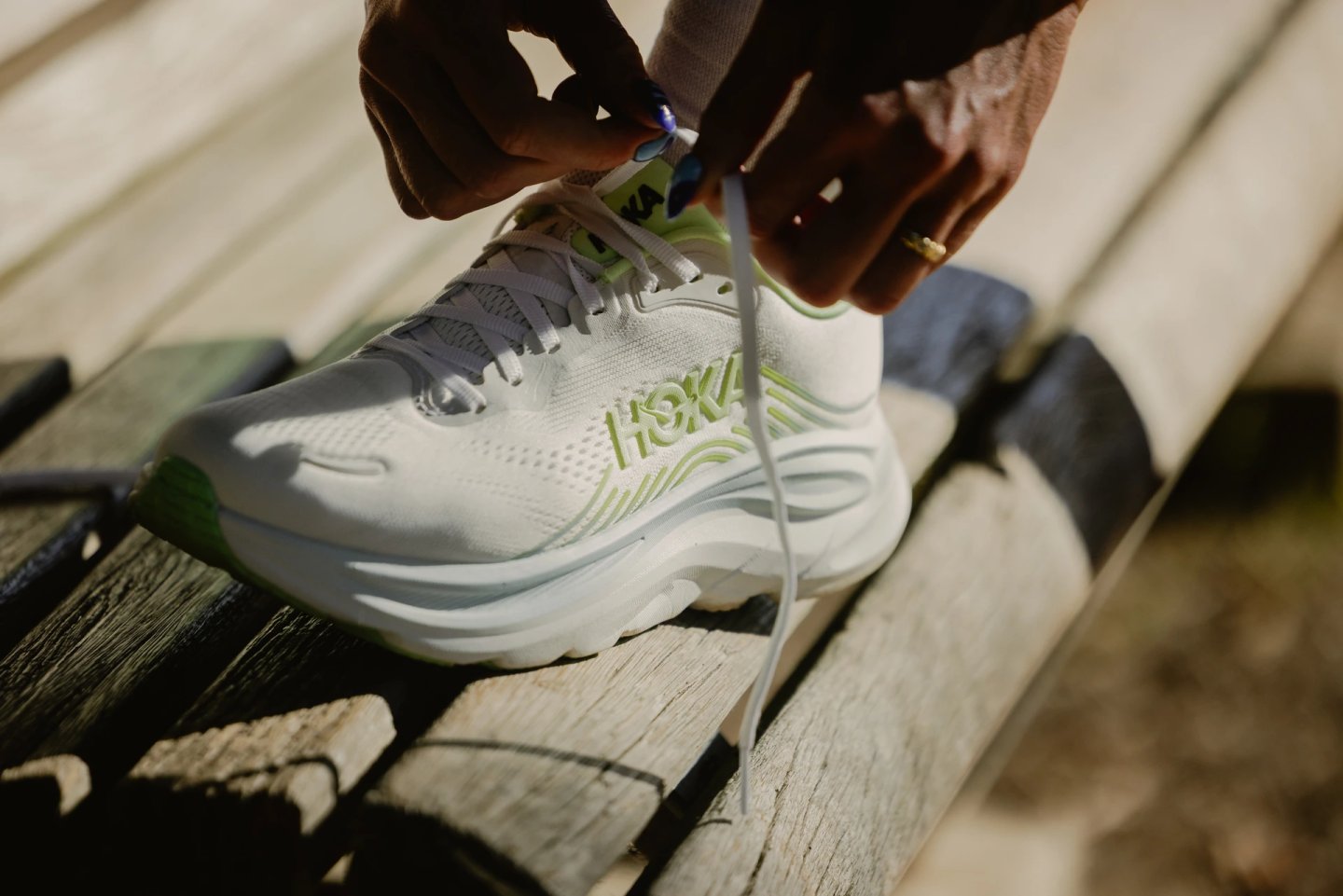 Close-up of hands tying laces on a white and lime green HOKA running shoe resting on a wooden bench outdoors.