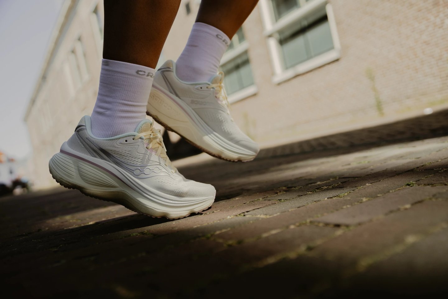 Close-up low-angle shot of white Saucony running shoes worn with white crew socks on an urban brick pavement.