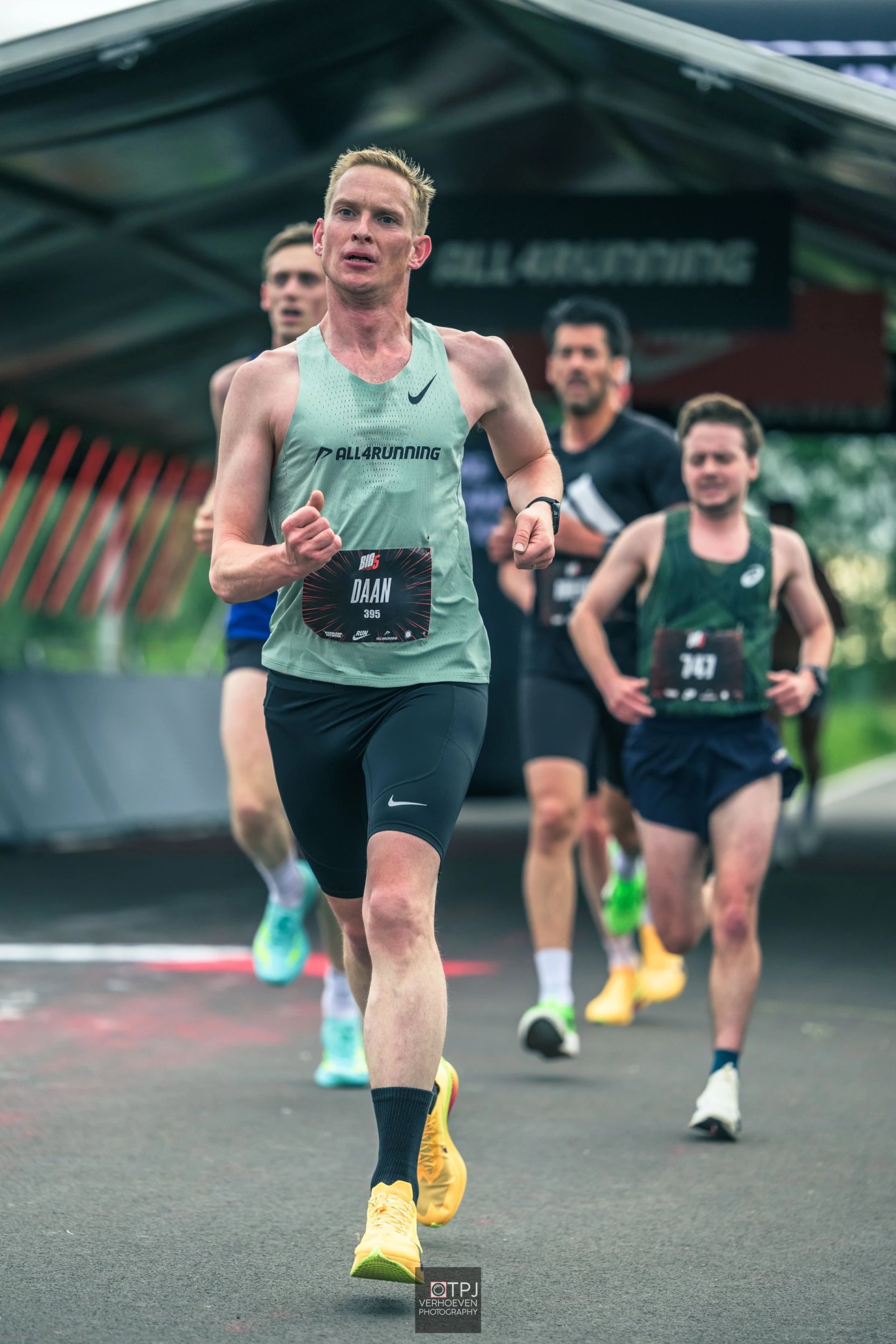 Male runner wearing green Nike singlet, black Nike tights, yellow racing shoes and black socks leading a road race, bib number 395.