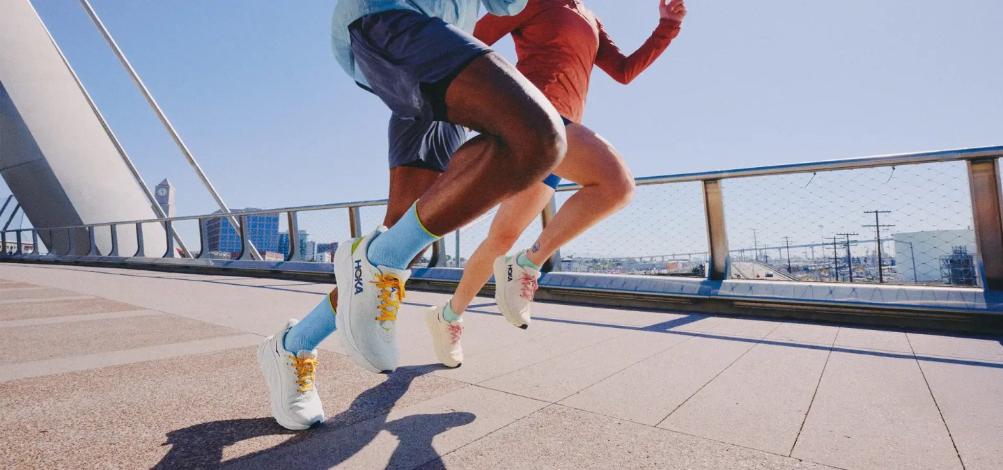 Two runners wearing white HOKA running shoes sprint across a sun-lit urban bridge with a city skyline behind them.