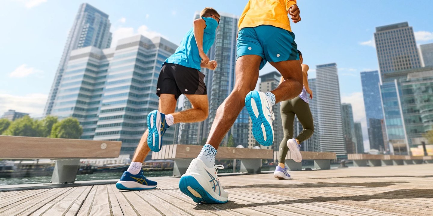 Three runners wearing Asics Cumulus 28 shoes sprint along a wooden boardwalk with a modern city skyline behind them.