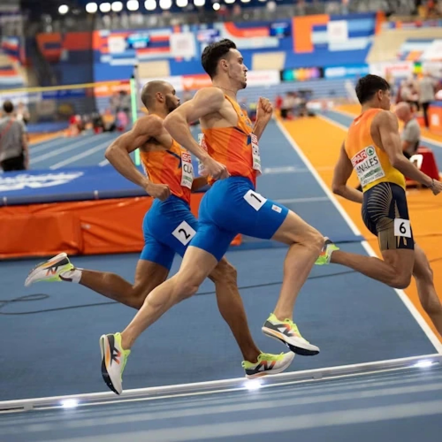 Three male sprinters racing on a blue indoor athletics track wearing orange uniforms and neon yellow running shoes.