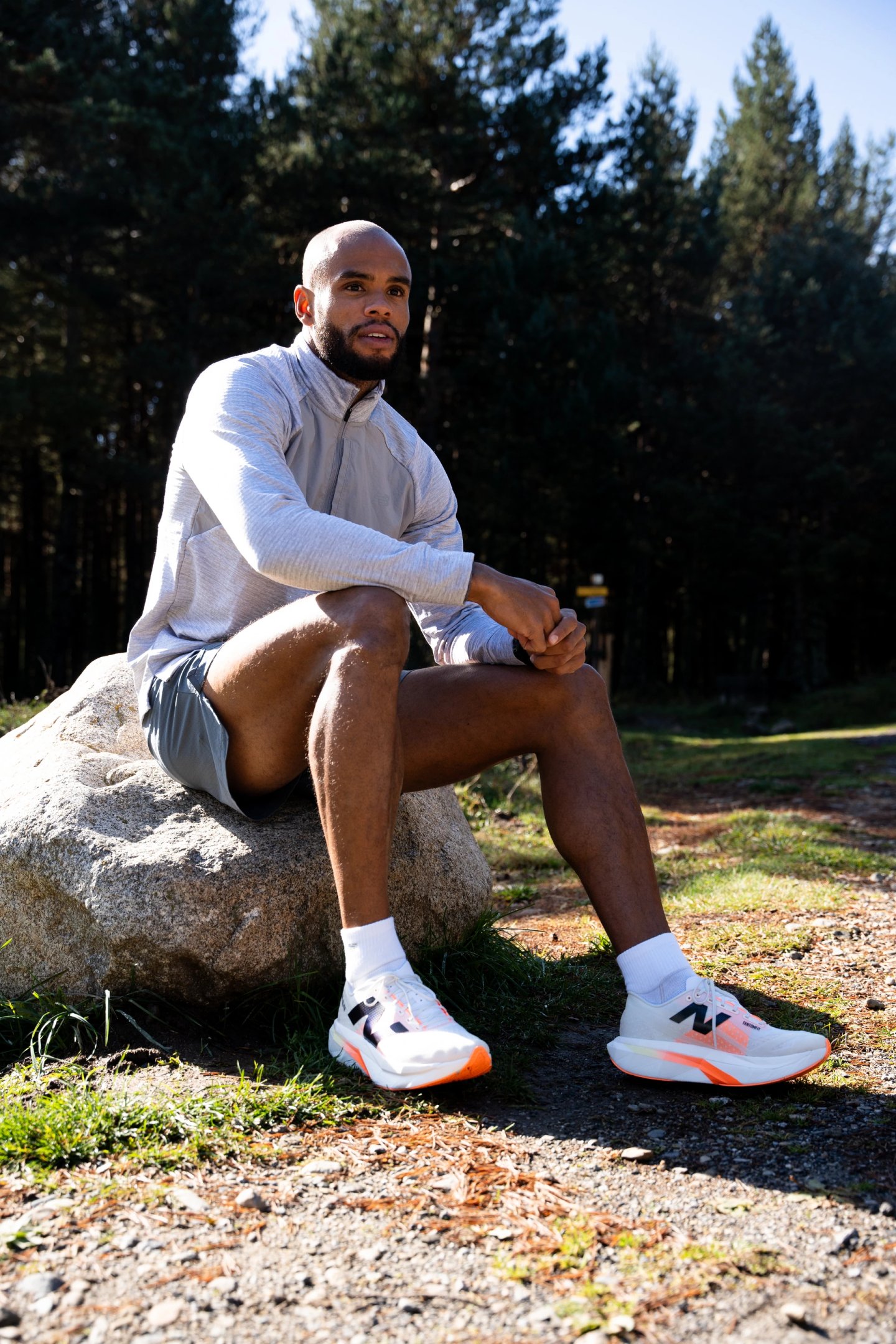 Male runner sitting on a large rock in a pine forest wearing white and orange New Balance running shoes and grey athletic wear.