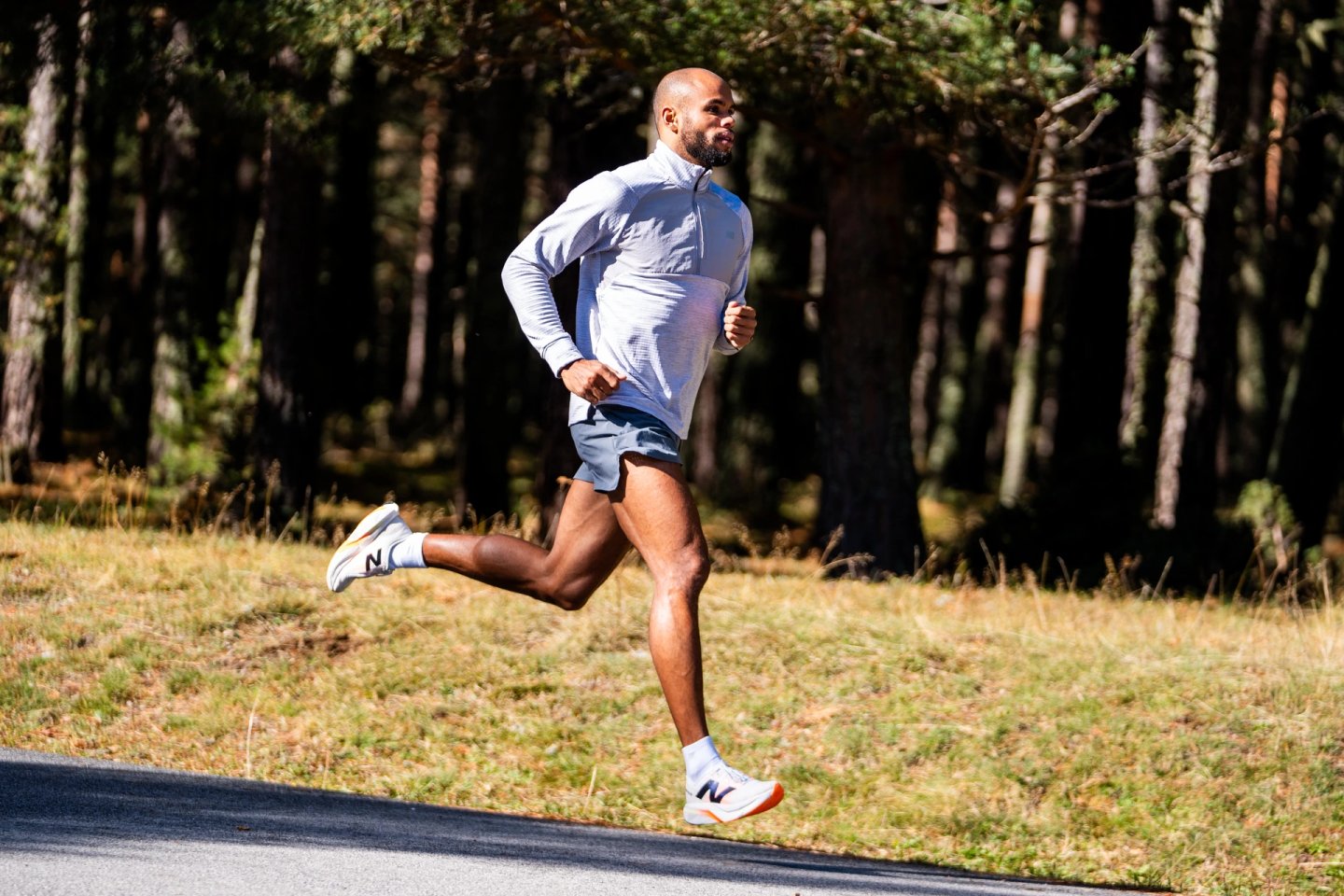 Male runner in grey half-zip top and blue shorts wearing white New Balance shoes with orange outsoles on a forest road.