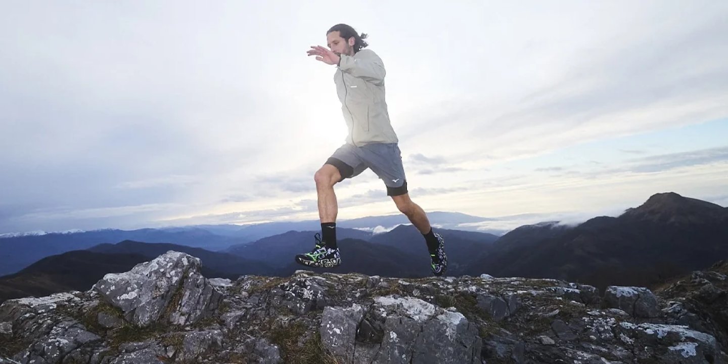 Male trail runner mid-leap across rocky mountain summit wearing grey jacket, shorts, and colourful trail shoes.