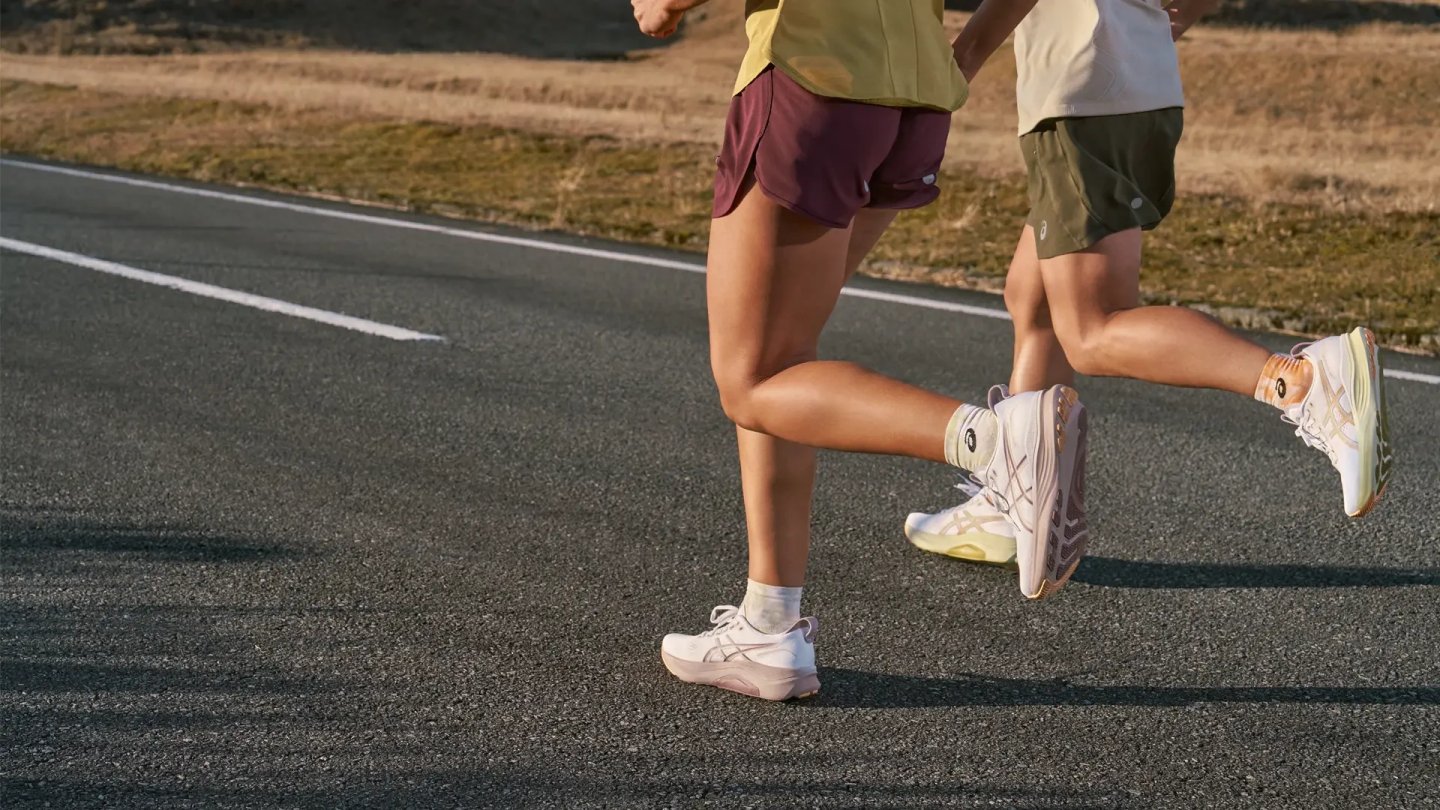 Two runners from the waist down wearing Asics Kayano 32 shoes in white and cream colorways on asphalt road