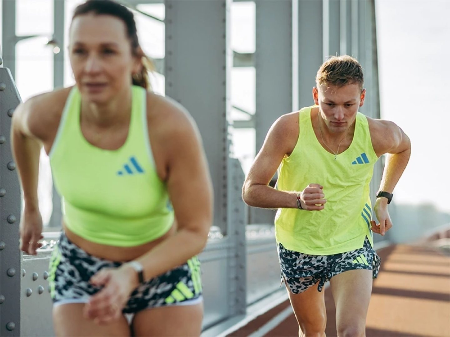 Man and woman running on a bridge wearing matching adidas neon yellow running tops and camo-print shorts