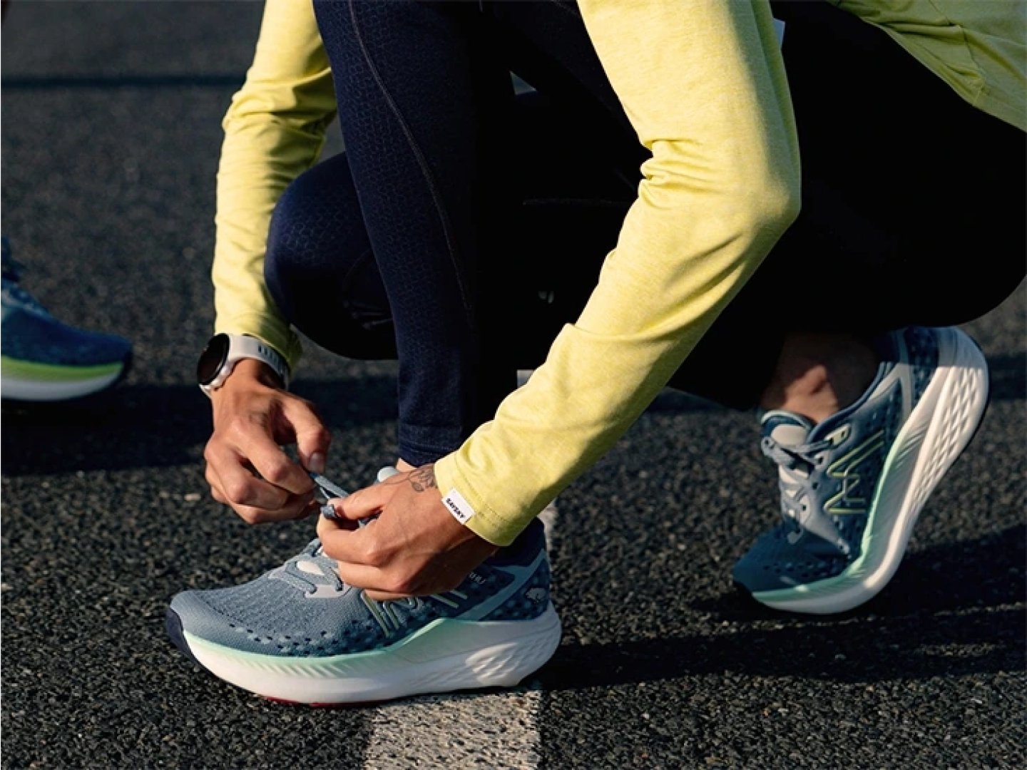 Runner in yellow top and navy tights tying laces on blue and white New Balance running shoes on asphalt road.
