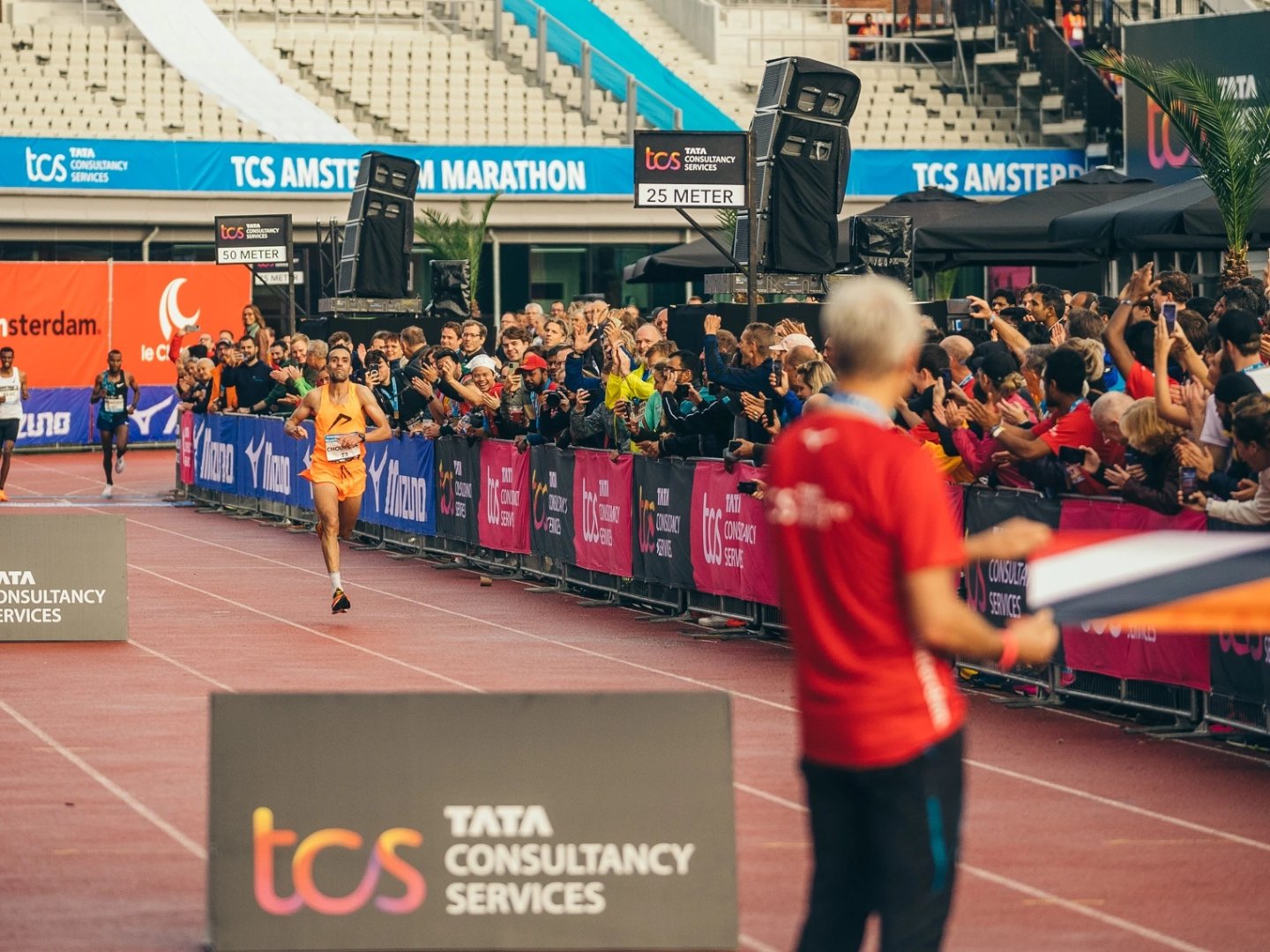 Elite male runner in orange kit approaches the finish line at TCS Amsterdam Marathon inside a stadium track, 25 metres to go.