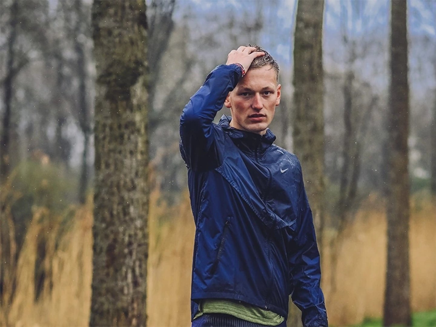 Male runner in a navy Nike running jacket standing in a rainy forest, hand on wet hair, looking at camera.