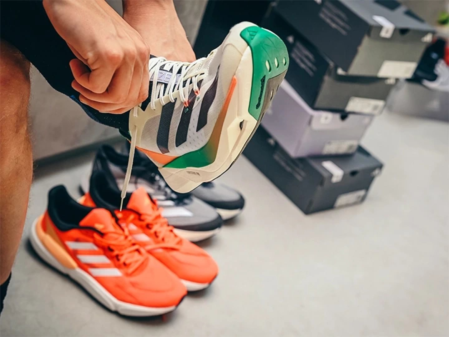 Person lacing up adidas running shoes in a store, with orange adidas trainers and grey runners on the floor nearby.