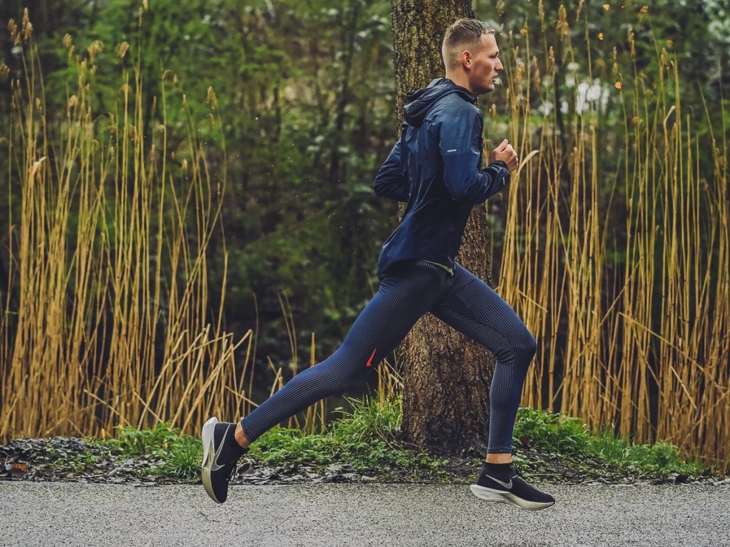 Male runner sprinting outdoors in black waterproof running shoes, navy tights and a blue hooded running jacket