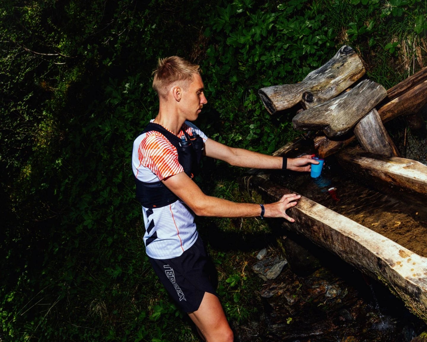 Male trail runner in white and orange jersey and black shorts refilling a blue soft flask at a wooden mountain trough