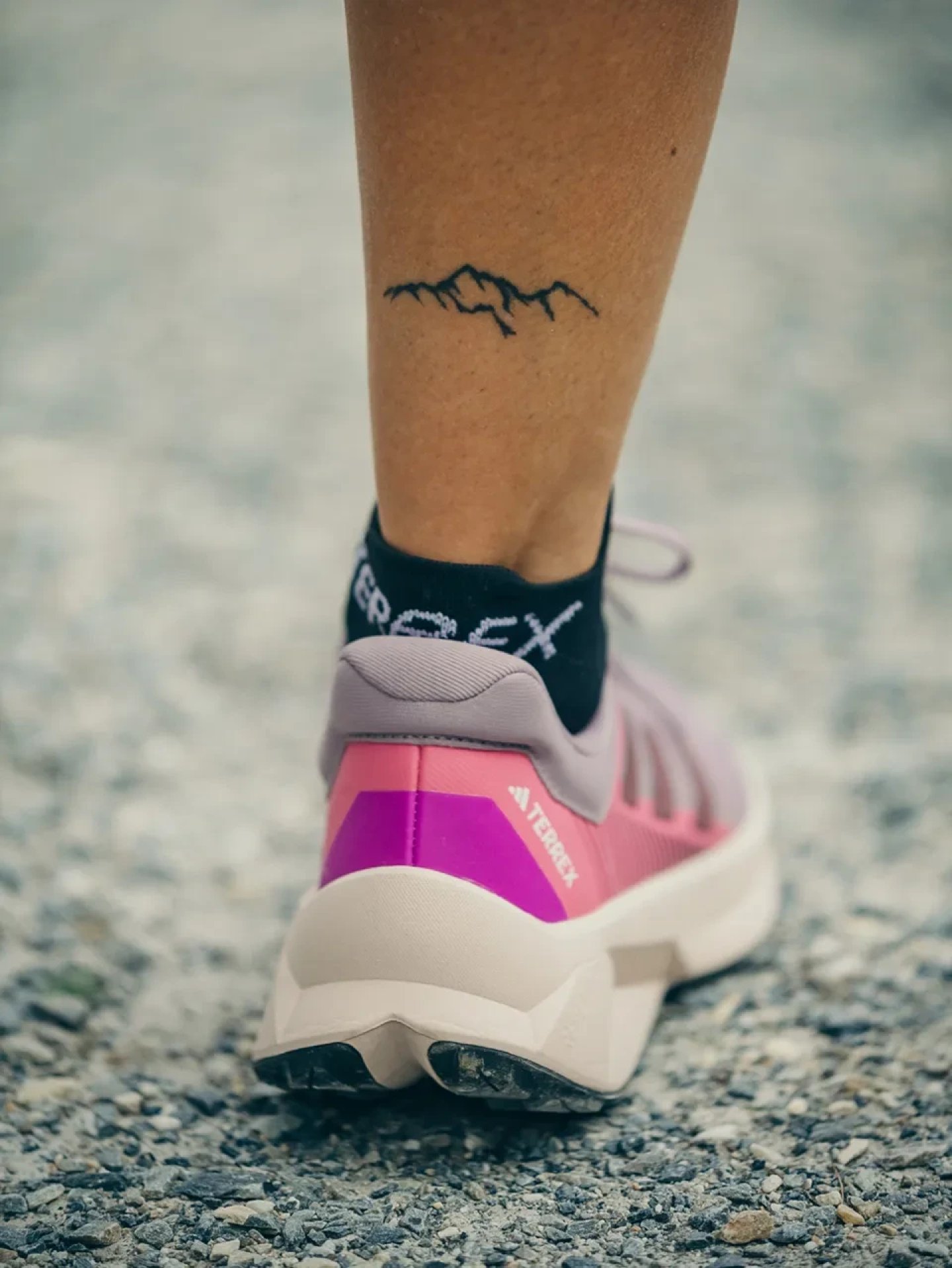 Rear view of an Adidas Terrex trail running shoe in pink and purple colorway worn with black Terrex ankle socks on gravel