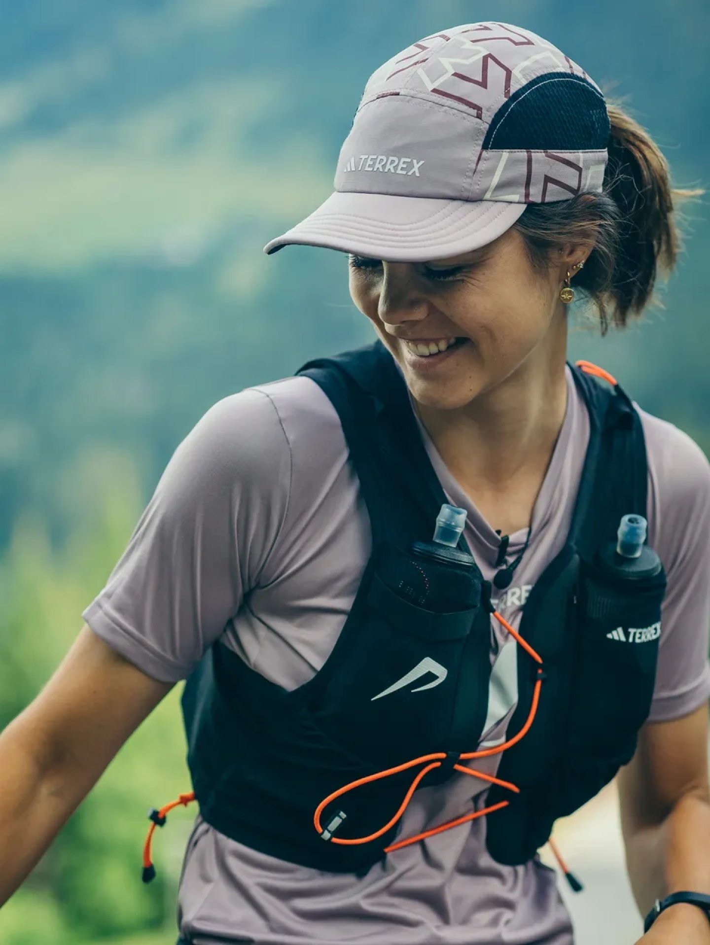 Smiling woman trail running wearing adidas Terrex hydration vest with soft flasks and adidas Terrex running cap in lilac.