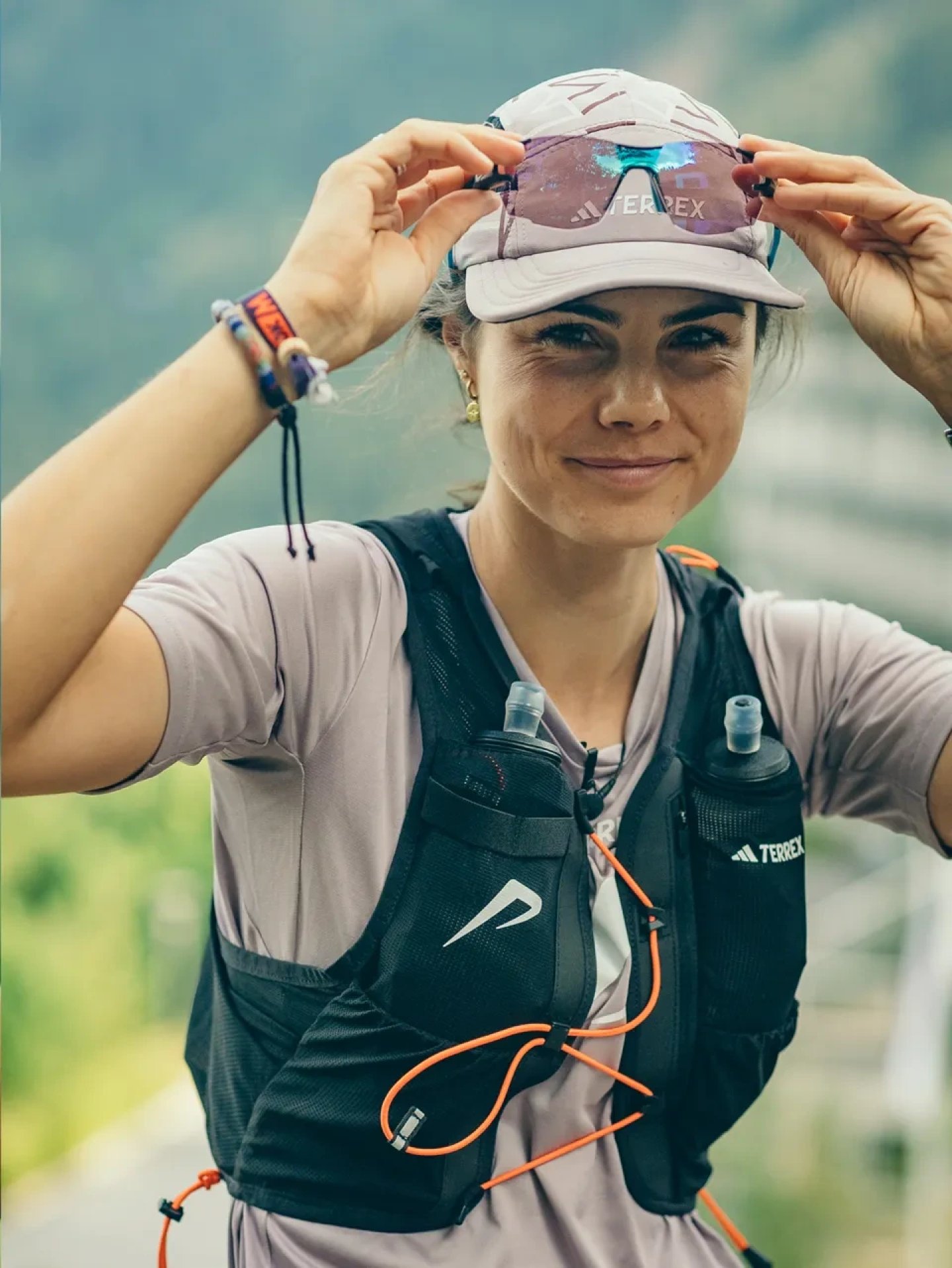 Female trail runner adjusting pink Adidas Terrex sunglasses, wearing black hydration vest with soft flasks and a running cap