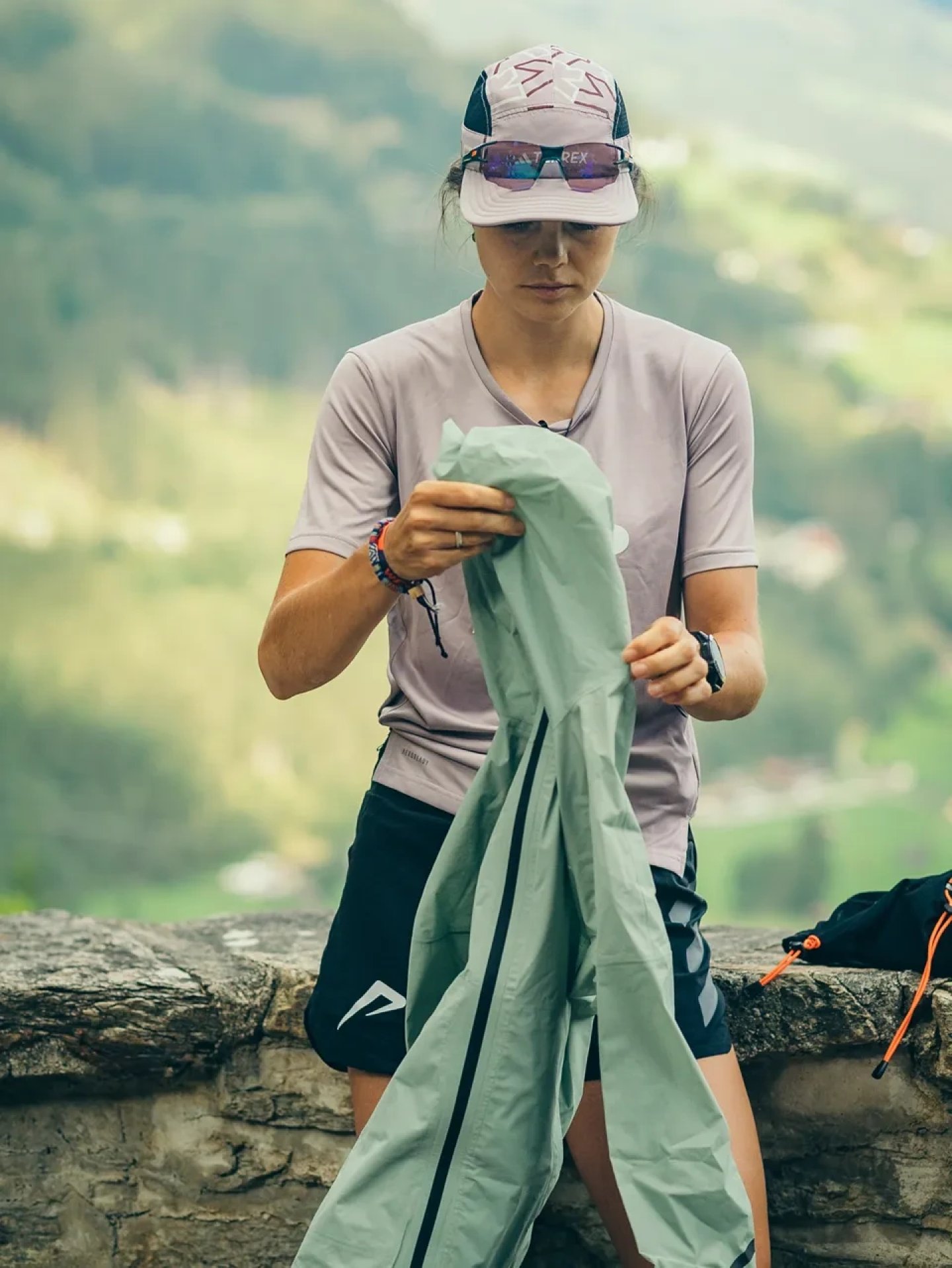 Woman in trail running cap, sunglasses and tee unfolding a lightweight mint green shell jacket on a stone wall in the mountains.