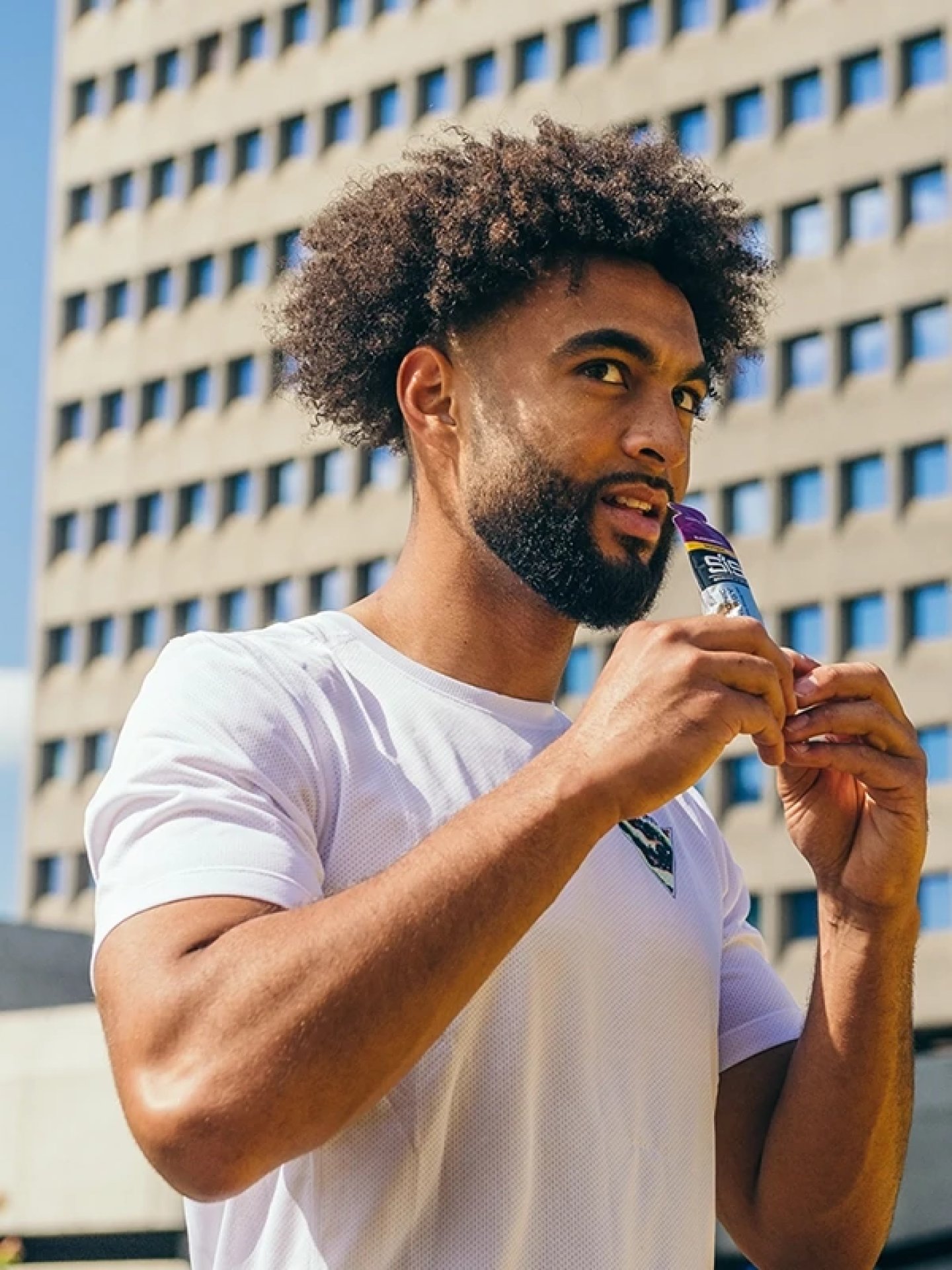Male runner in white mesh t-shirt consuming a SiS energy gel sachet outdoors in an urban setting