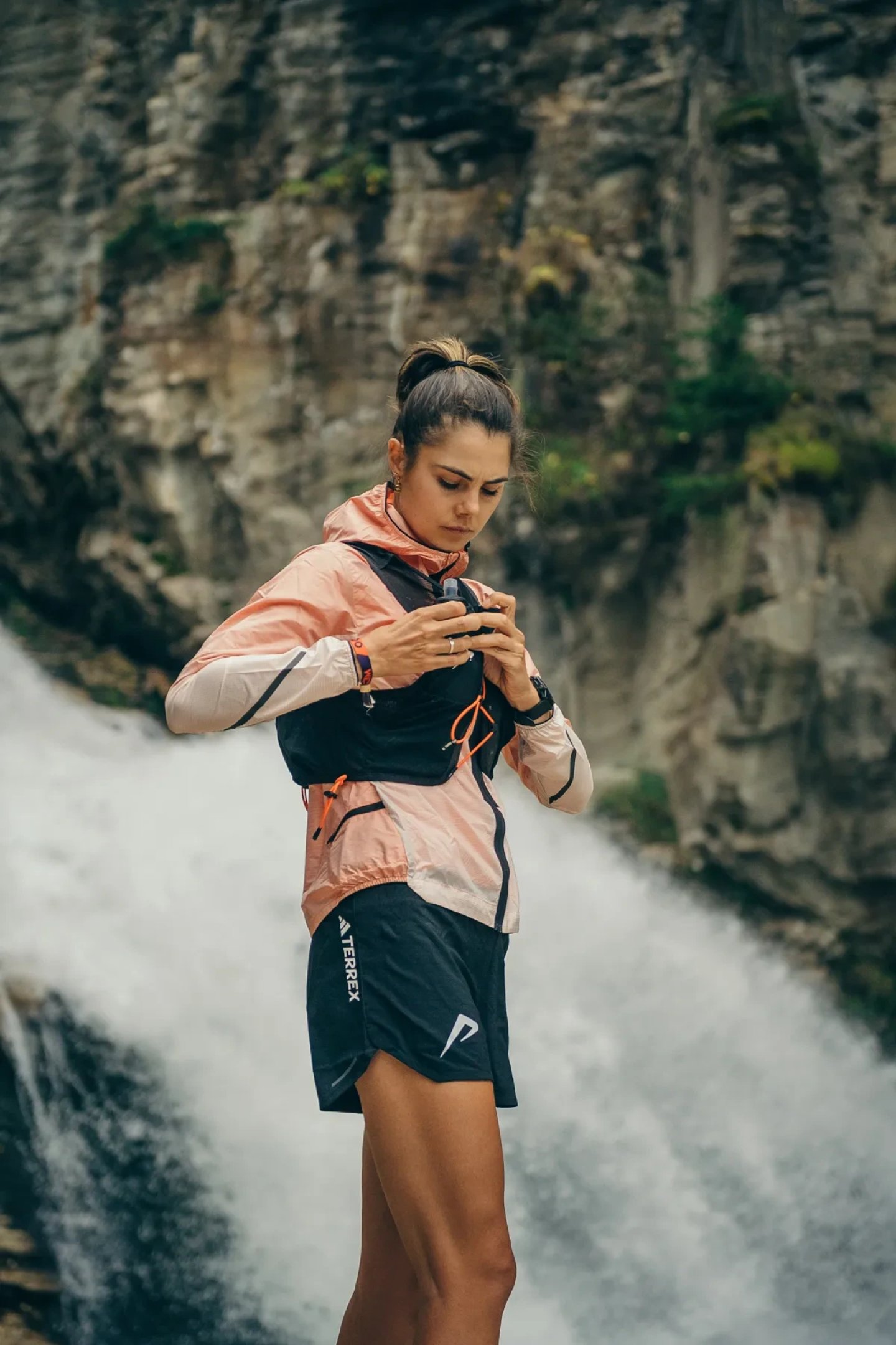 Woman in adidas Terrex trail running shorts, pink windproof jacket and black hydration vest adjusting fit near a waterfall