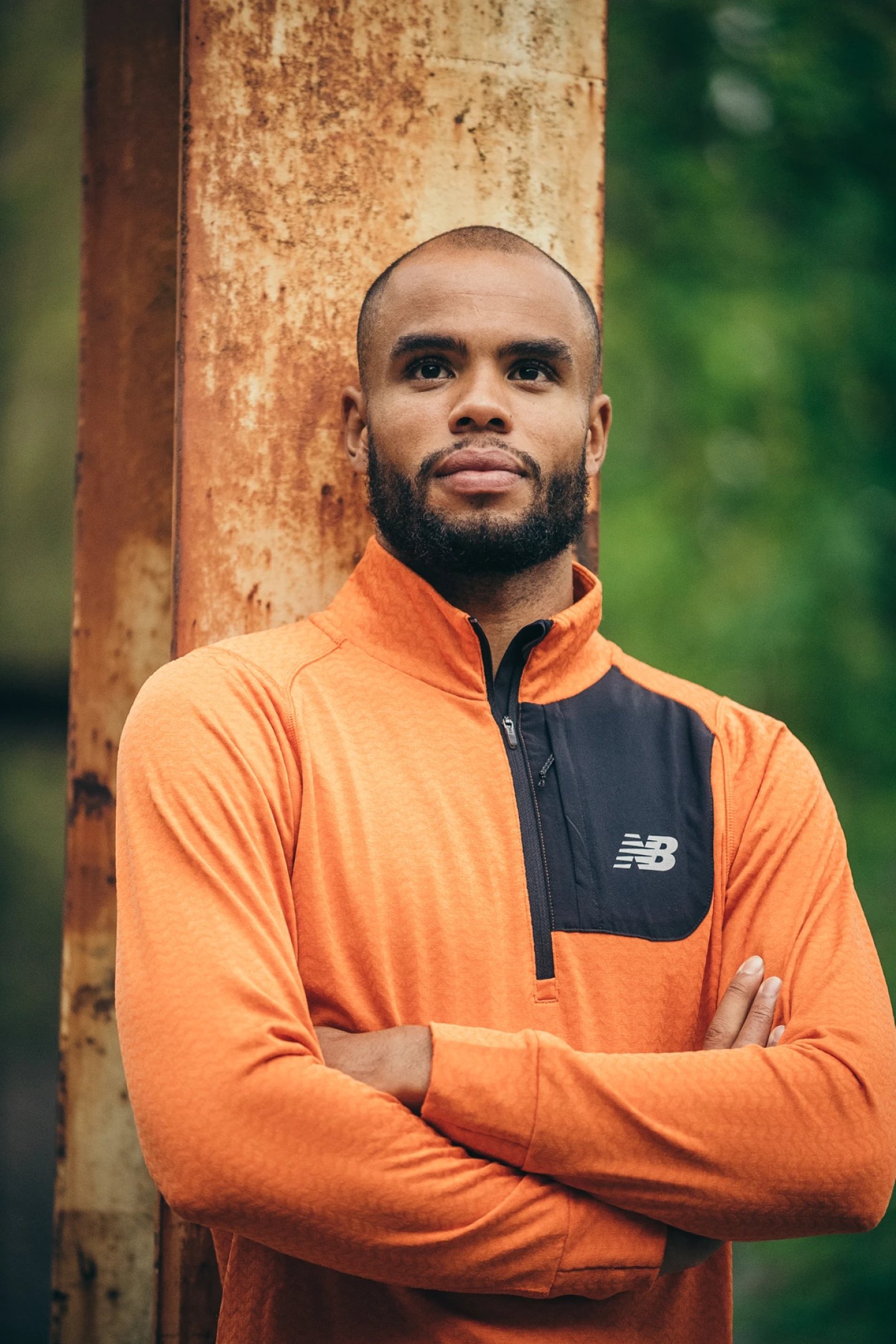 Man wearing a New Balance orange and black quarter-zip running top, arms crossed, standing outdoors against a rustic post.