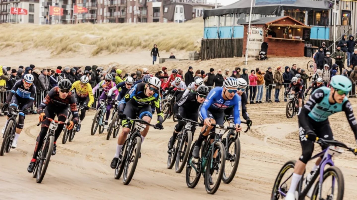 Peloton of mountain bikers racing on a sandy beach at Combiklassement Egmond, with spectators watching from barriers.