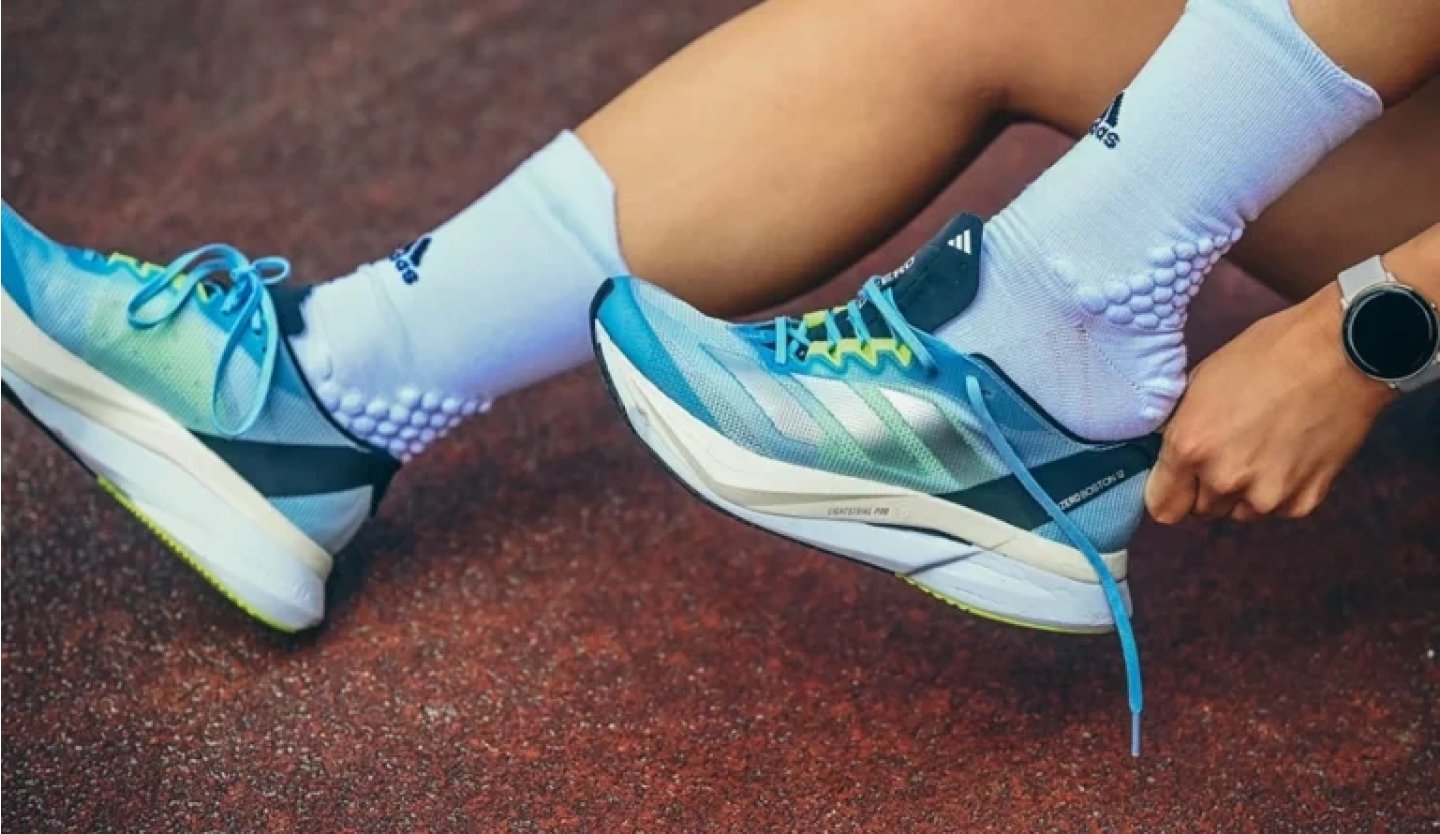 Close-up of adidas blue and teal running shoes being laced by a runner wearing white grip socks on a red track