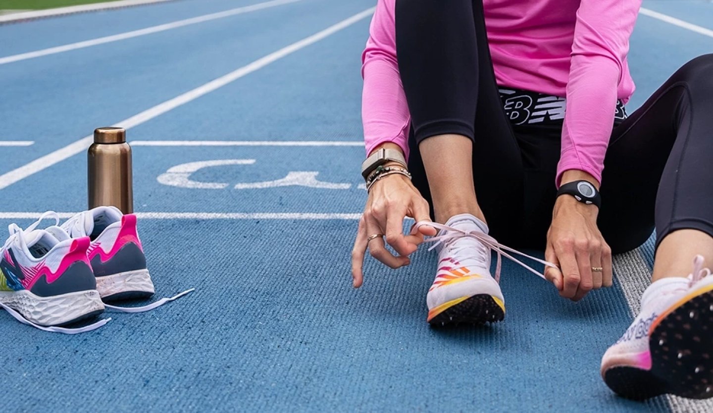 Woman in pink top and black leggings tying laces on multicolour racing spikes while seated on a blue running track.