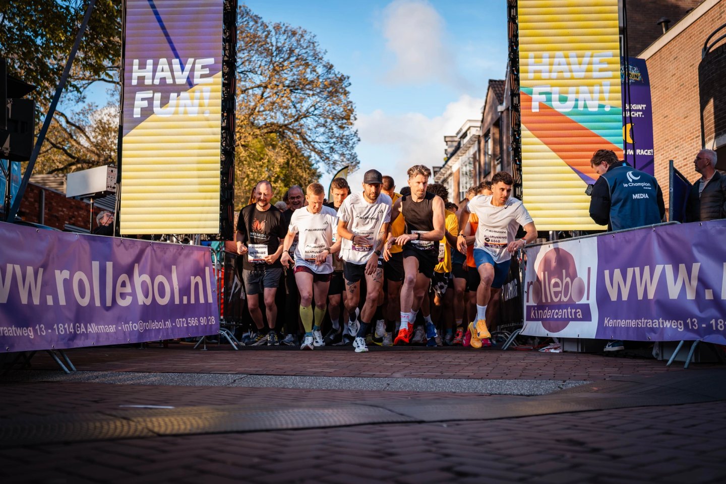 Group of runners crossing the start line at the Alkmaar City Run event, passing under large LED screens displaying 'Have Fun'.