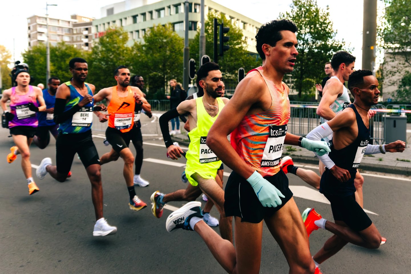 Elite male runners in a close pack racing through city streets at the TCS Amsterdam Marathon, wearing race bibs and performance kit.