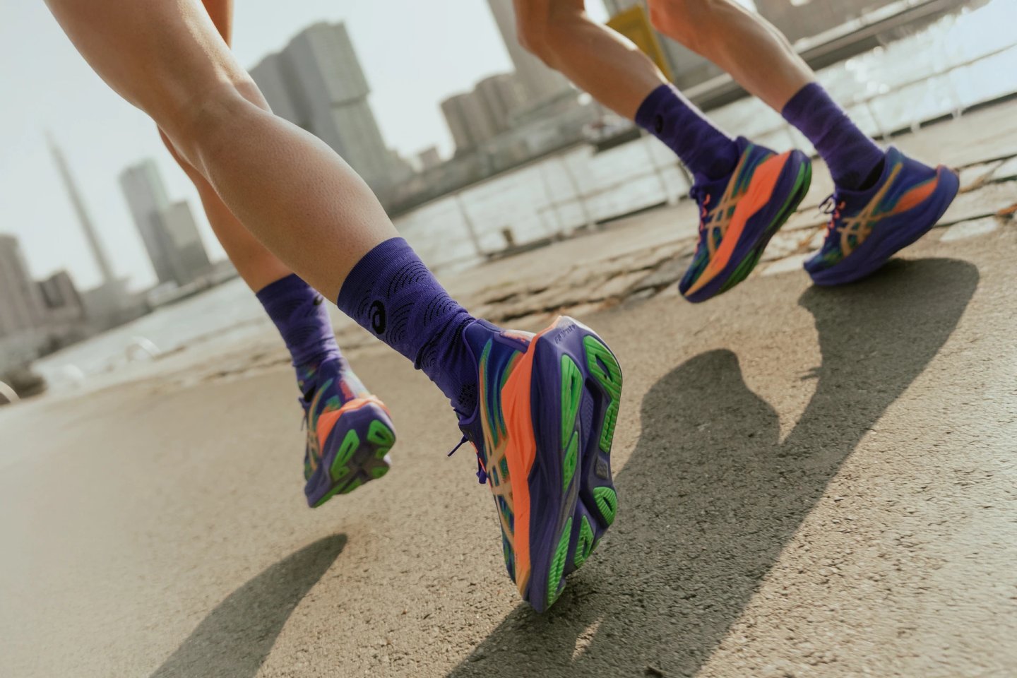 Close-up of two runners wearing purple and orange ASICS running shoes and purple ASICS socks on a concrete urban running course.