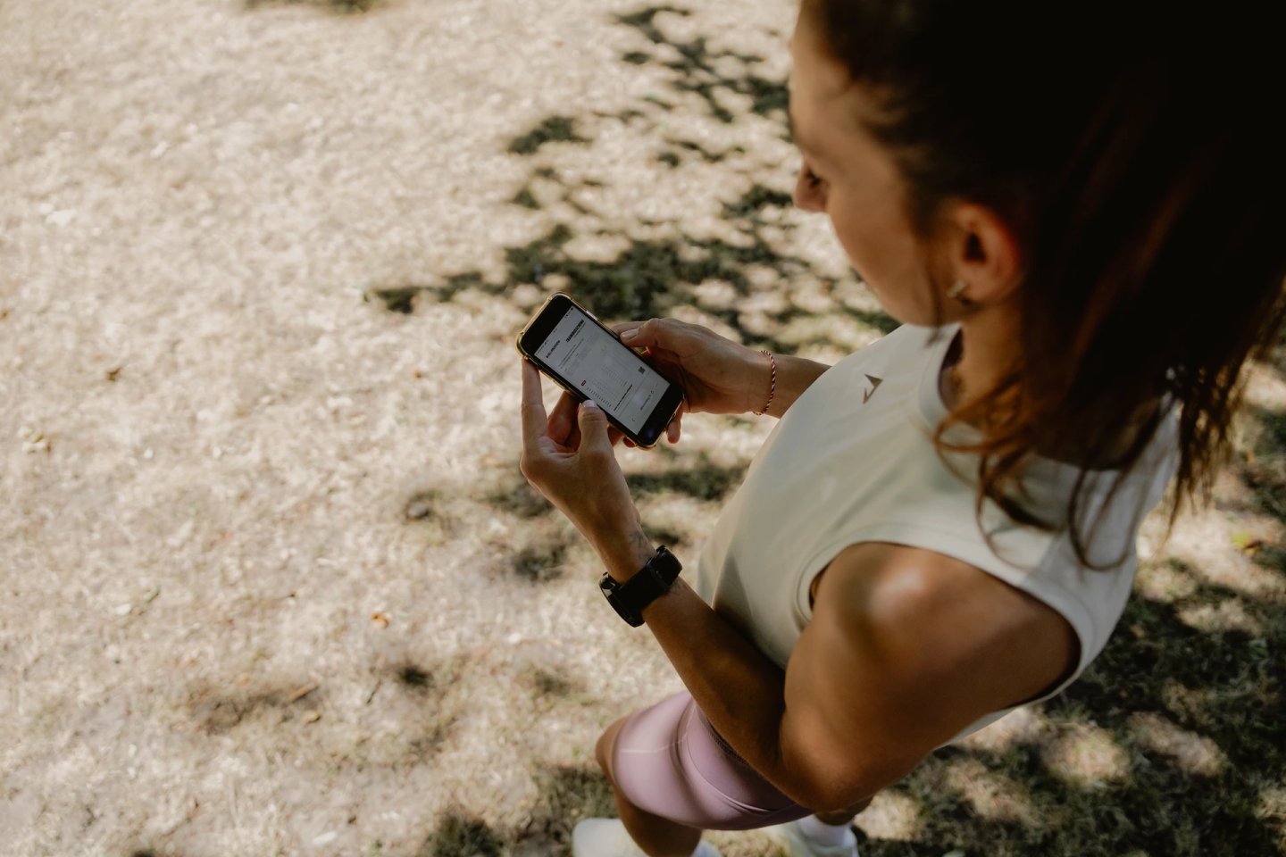 Female runner in white vest and pink leggings checking smartphone with GPS running watch on wrist, outdoors on sunny day.