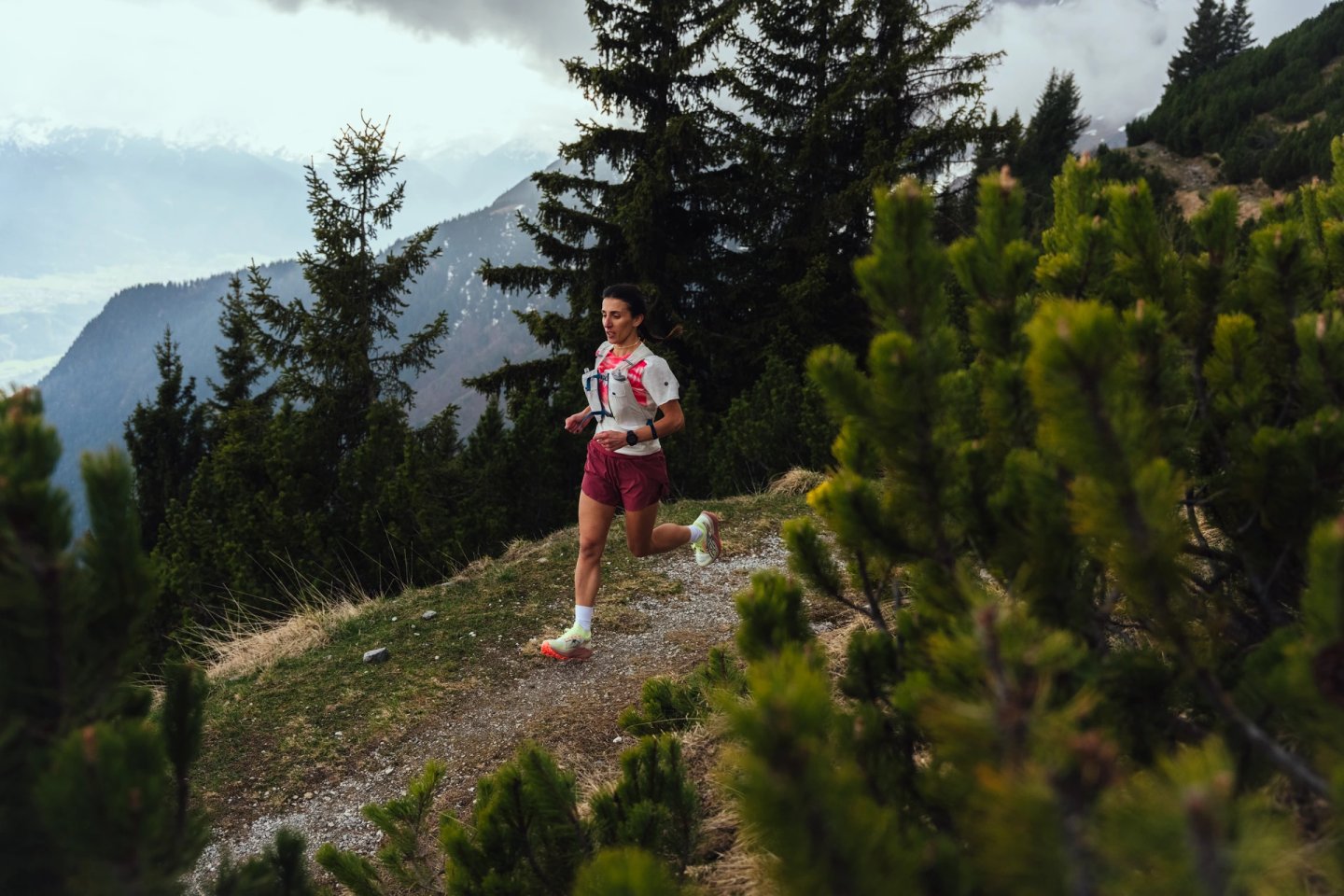Female trail runner in white top, burgundy shorts and neon trail shoes running on a rocky alpine path with mountains behind.