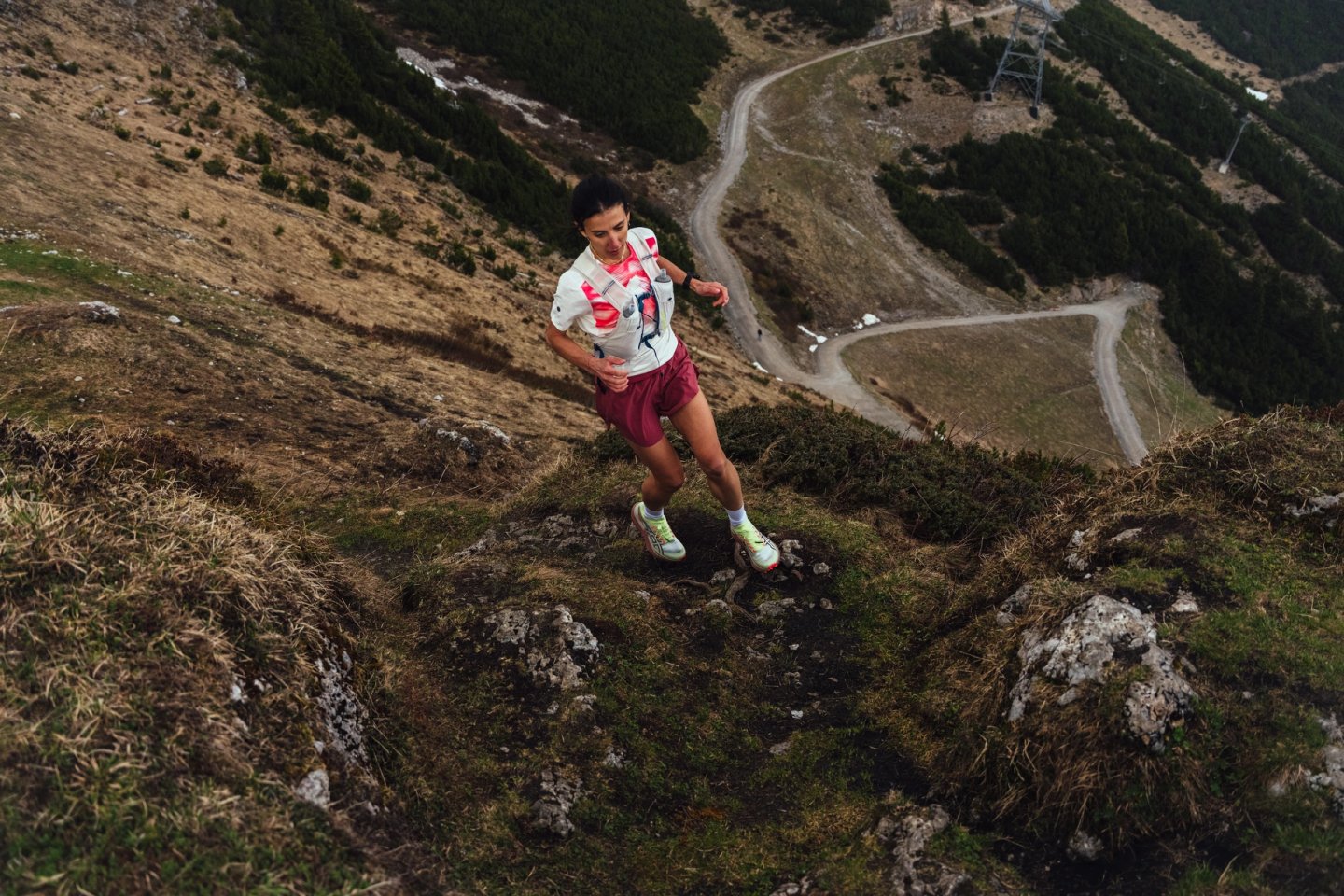 Aerial view of a female trail runner descending a rocky mountain trail wearing a white vest, burgundy shorts, and neon trail shoes.