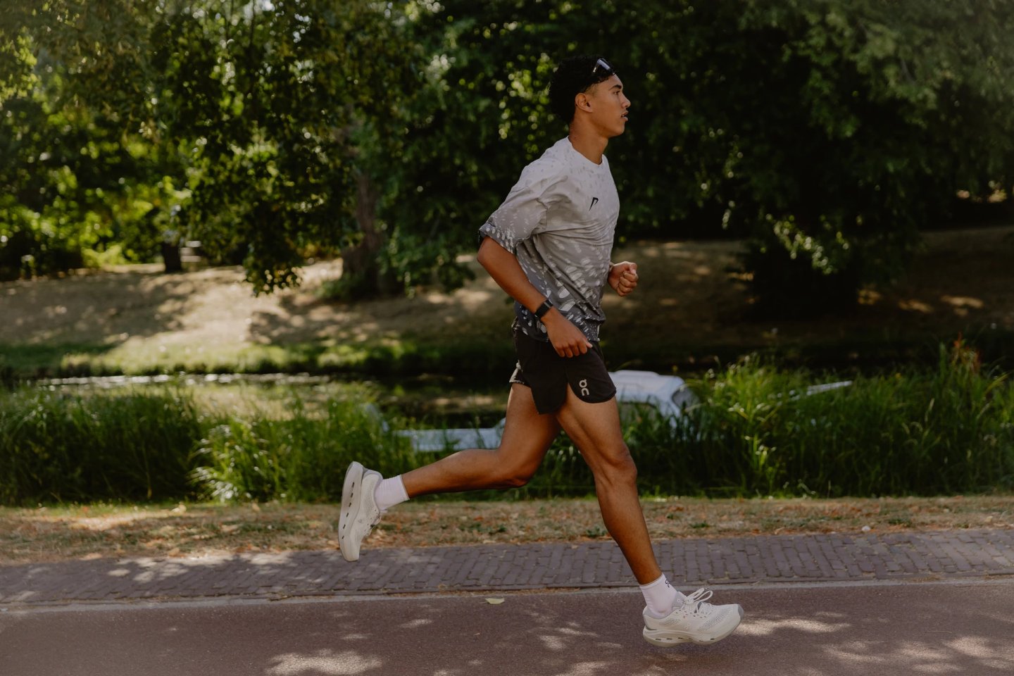 Man running outdoors in white patterned t-shirt, black On shorts, white socks and white On running shoes on a park path.