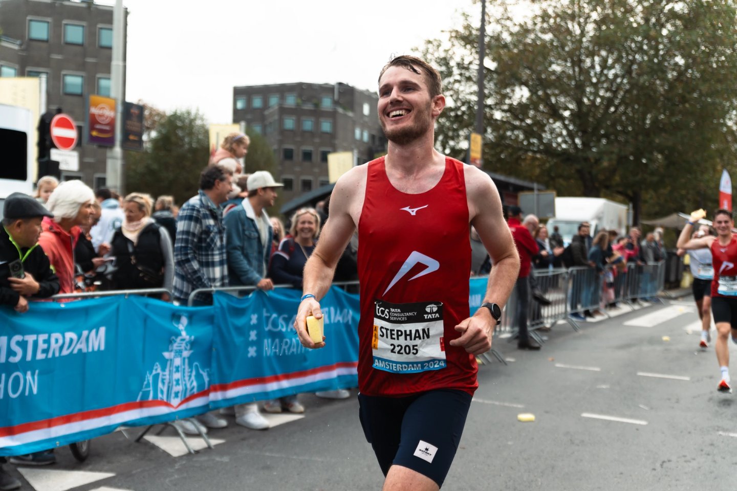 Smiling male runner in red Mizuno singlet and dark compression shorts racing at TCS Amsterdam Marathon 2024, bib number 2205.