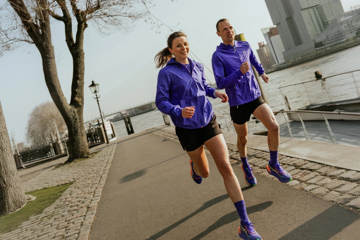 Man and woman running together on a cobblestone riverside path in Rotterdam, both wearing purple running jackets and black shorts.