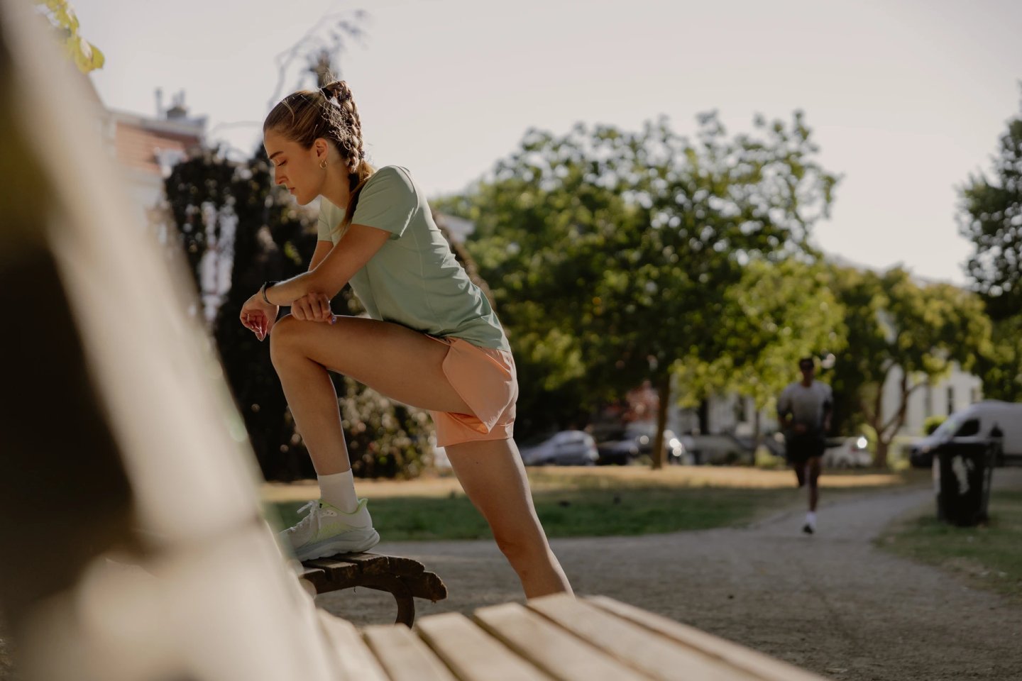 Woman in green running t-shirt and peach shorts resting one foot on a park bench while checking her smartwatch outdoors.