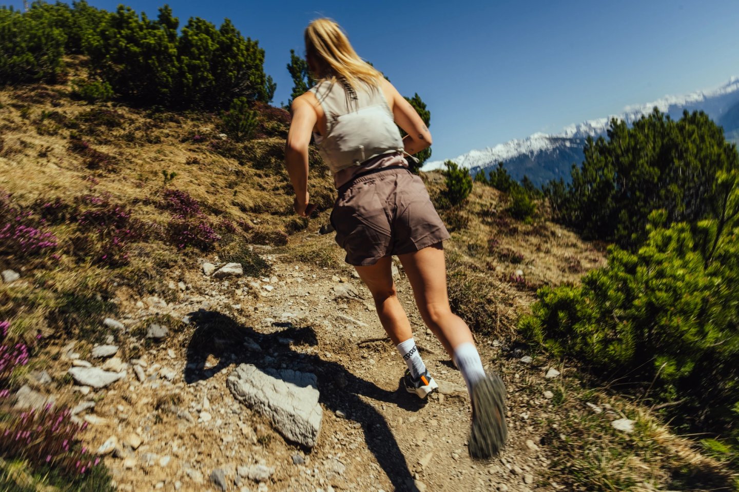 Woman trail running uphill on a rocky alpine path wearing shorts, tank top, white crew socks and trail shoes