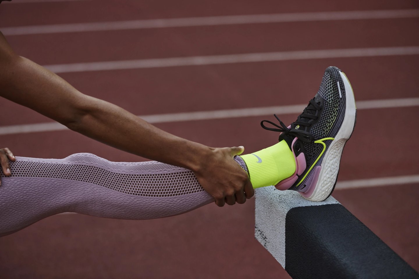 Close-up of Nike Invincible running shoe in black and pink on a red athletics track, worn with a neon yellow Nike sock