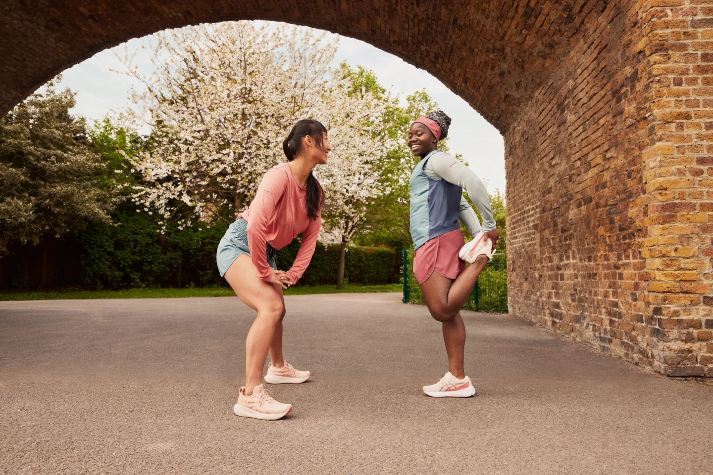 Two women in ASICS running shoes and apparel stretching together under a brick arch in a park in spring