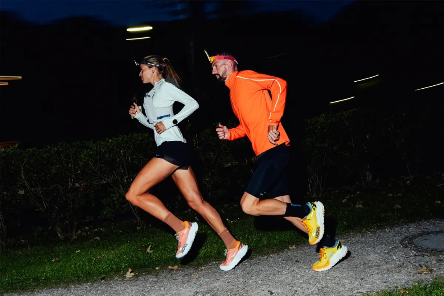 Man and woman running together outdoors at night, wearing headlamps and bright running apparel including an orange jacket.