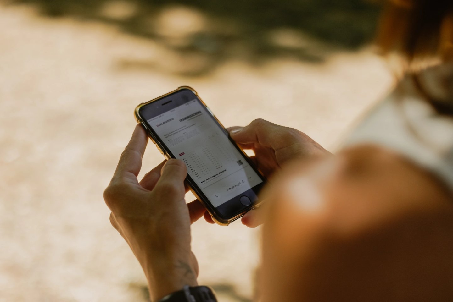Person holding a smartphone with both hands, browsing a running gear website outdoors in warm sunlight.