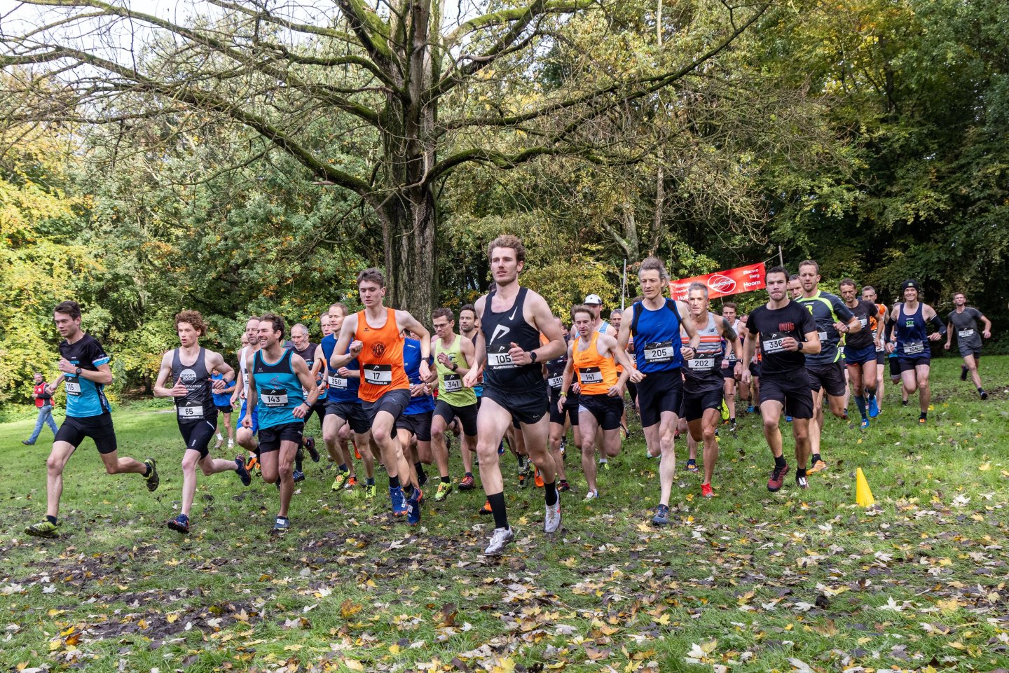 Large group of runners racing across a muddy grass park course in autumn, led by Menno Munster wearing bib 105.