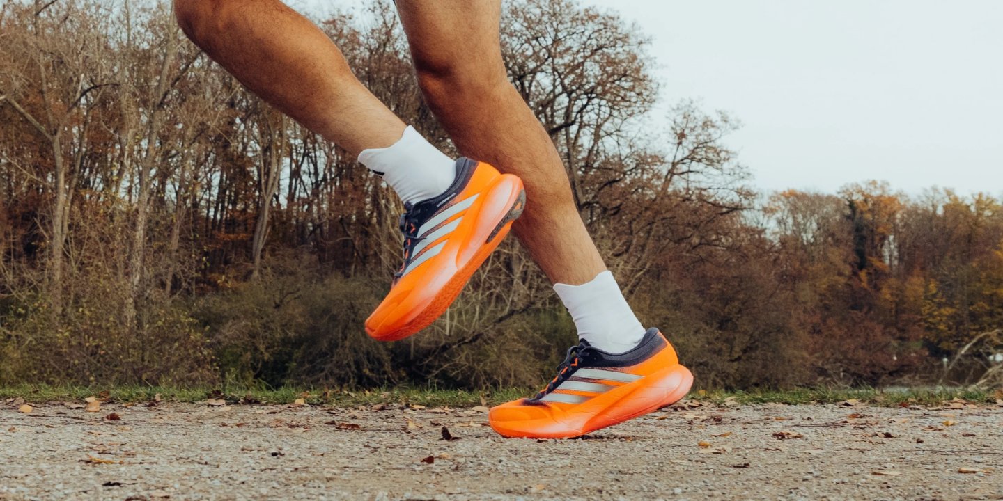 Male runner mid-stride wearing bright orange Adidas running shoes with navy upper and silver Three Stripes on autumn road