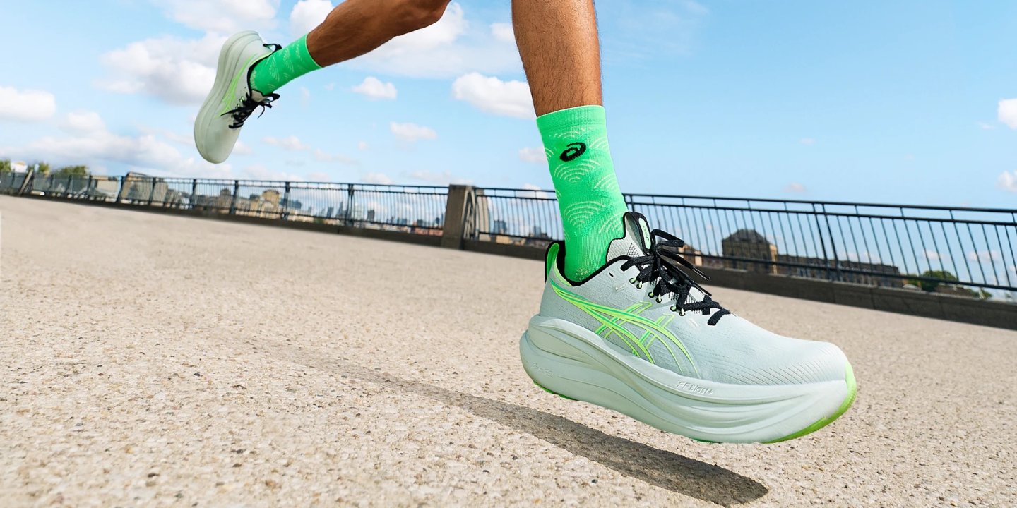 Male runner mid-stride wearing ASICS Gel-Nimbus 28 shoes in mint and neon green with matching ASICS socks on an urban bridge
