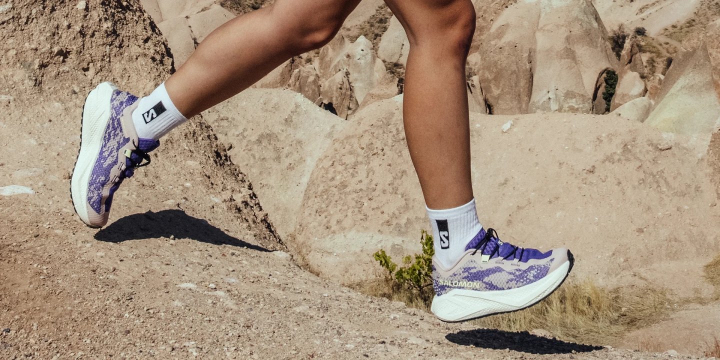 Runner in Salomon Aeroglide GRVL4 shoes in purple and white, mid-stride on rocky desert trail with sandstone formations.