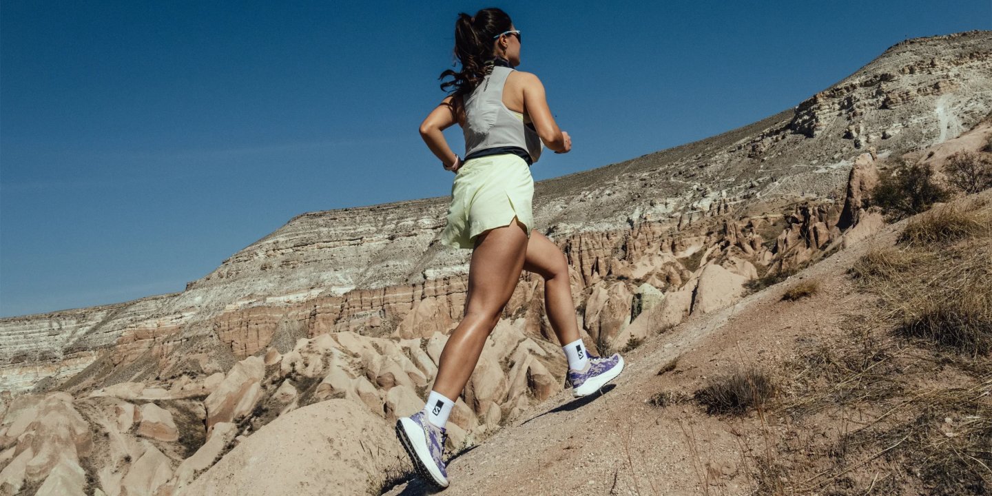 Female trail runner in Salomon shoes and lime shorts running uphill through a rocky desert canyon.